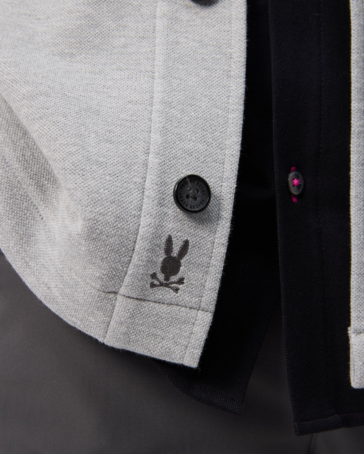 A close-up of the Psycho Bunny BEDFORD SHIRT JACKET - B6J290B200 in gray fabric highlights a rabbit head and crossbones logo, with black and dark red buttons against a dark background.