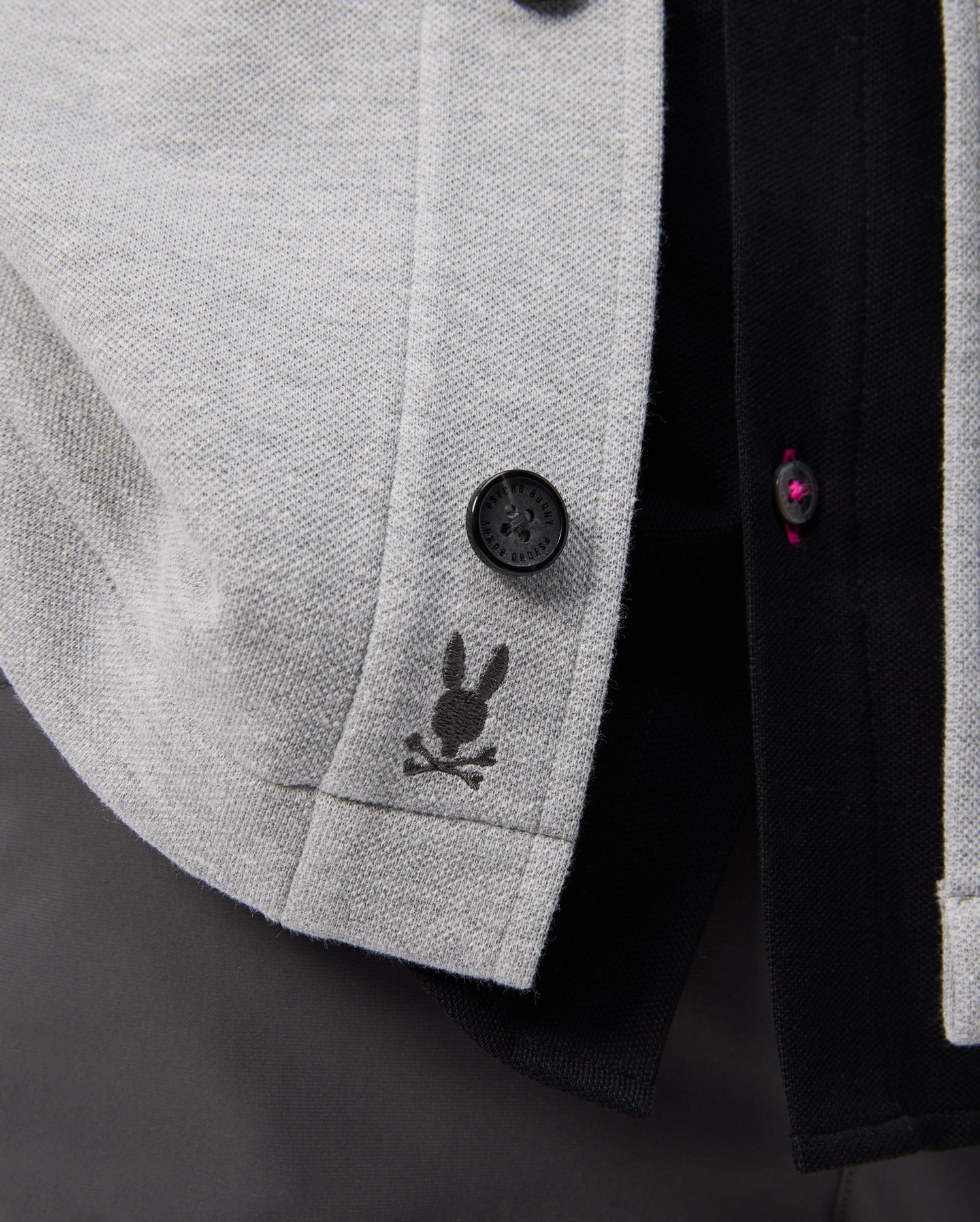 A close-up of the Psycho Bunny BEDFORD SHIRT JACKET - B6J290B200 in gray fabric highlights a rabbit head and crossbones logo, with black and dark red buttons against a dark background.