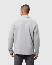 A person with short hair, seen from behind, wearing a light gray Psycho Bunny BEDFORD SHIRT JACKET - B6J290B200 with buttoned cuffs and dark gray pants, standing against a light gray background.