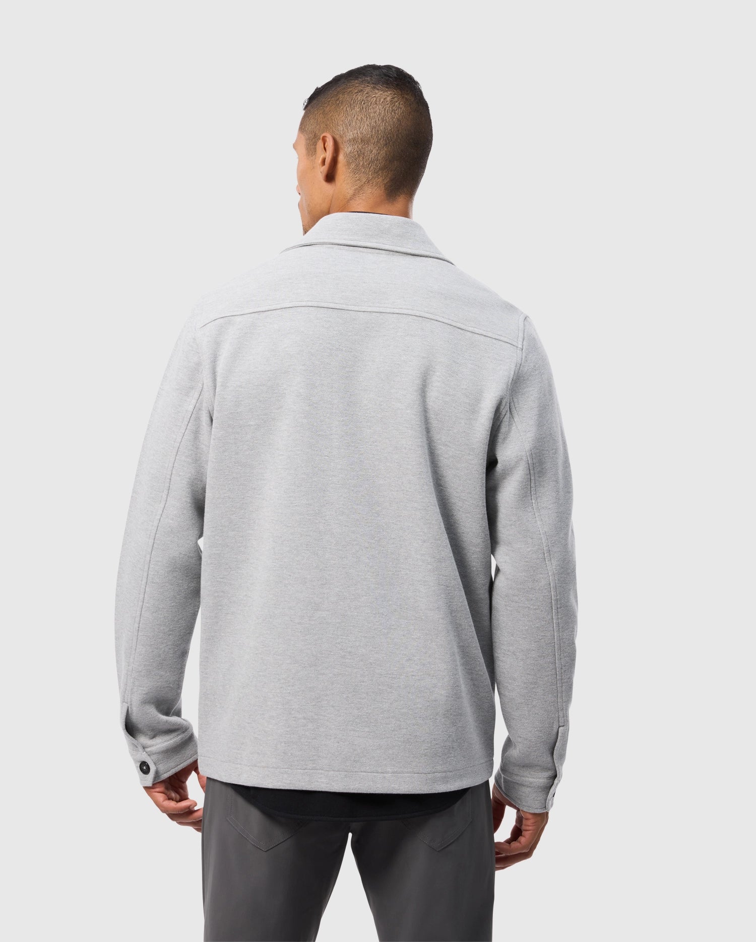 A person with short hair, seen from behind, wearing a light gray Psycho Bunny BEDFORD SHIRT JACKET - B6J290B200 with buttoned cuffs and dark gray pants, standing against a light gray background.