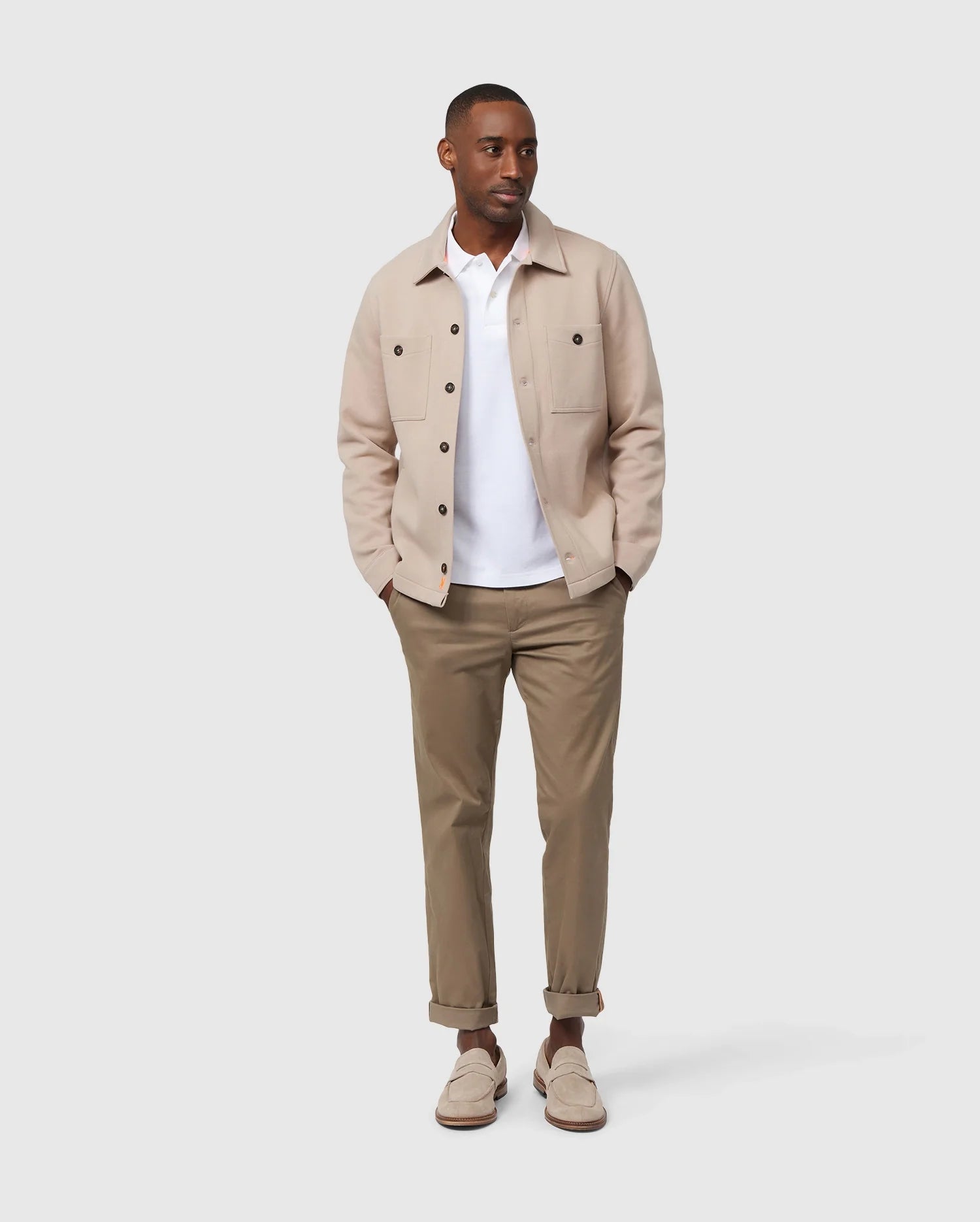 A man wears a beige Psycho Bunny MENS BEDFORD SHIRT JACKET - B6J290B200, featuring logo-engraved buttons, over a white shirt and tan pants as he stands with his hands in his pockets. This stylish, casual outfit is ideal for a smart-casual occasion. The simple background highlights the ensemble's sophisticated details.