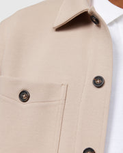 Close-up of the Psycho Bunny MENS BEDFORD SHIRT JACKET - B6J290B200 in beige, showcasing logo-engraved dark buttons and work-jacket style pockets, layered over a white shirt. The textured fabric and focus on the upper part highlight its structured design akin to a classic Bedford shirt jacket.