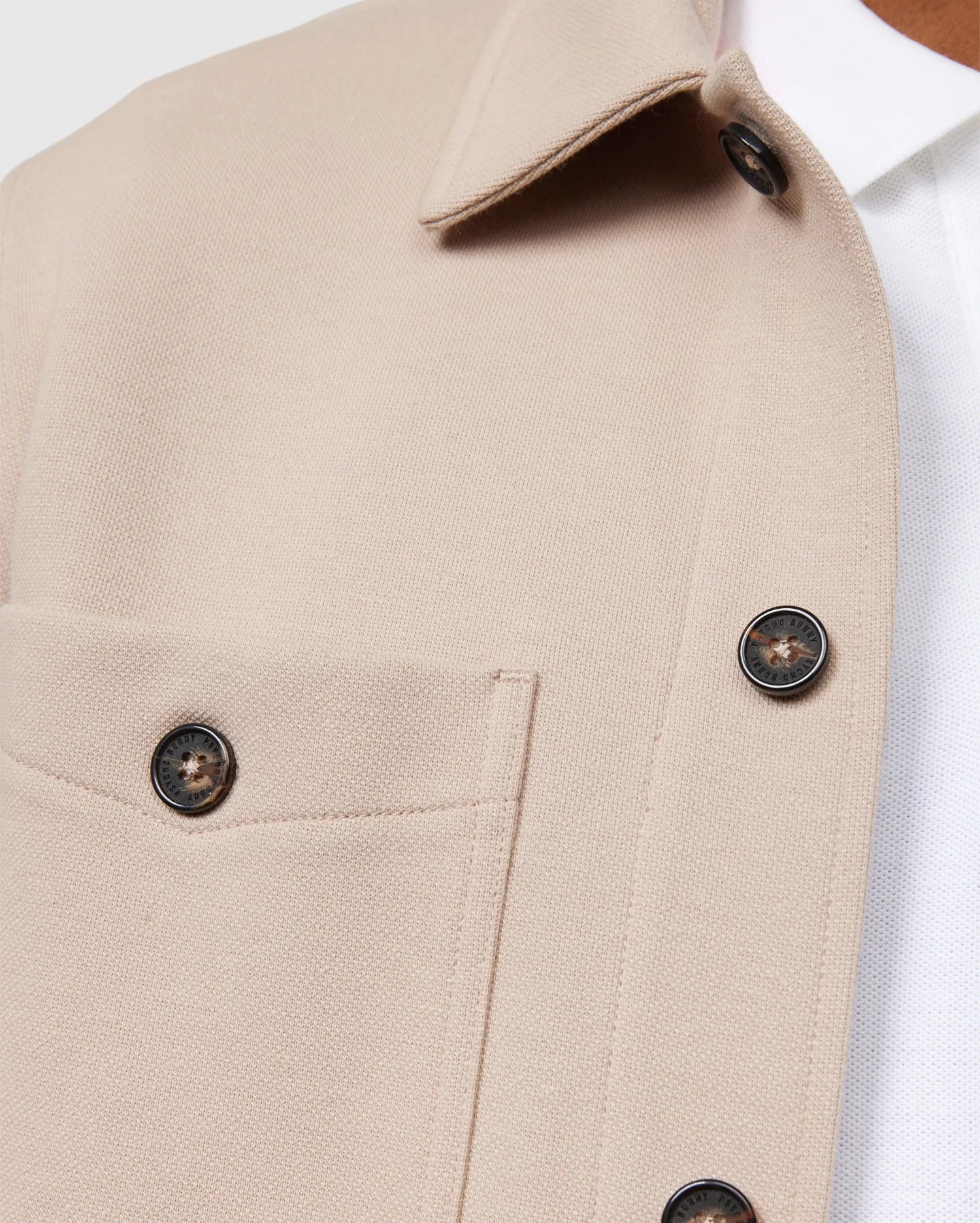 Close-up of the Psycho Bunny MENS BEDFORD SHIRT JACKET - B6J290B200 in beige, showcasing logo-engraved dark buttons and work-jacket style pockets, layered over a white shirt. The textured fabric and focus on the upper part highlight its structured design akin to a classic Bedford shirt jacket.
