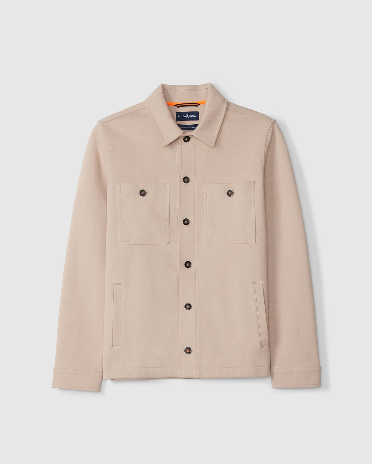 A beige MENS BEDFORD SHIRT JACKET - B6J290B200 from Psycho Bunny, featuring a collar and two work-jacket style pockets, displayed on a plain white background. The jacket is adorned with logo-engraved black buttons and boasts a sleek design, complete with long sleeves and an interior tag.