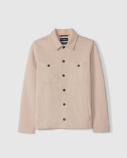 A beige MENS BEDFORD SHIRT JACKET - B6J290B200 from Psycho Bunny, featuring a collar and two work-jacket style pockets, displayed on a plain white background. The jacket is adorned with logo-engraved black buttons and boasts a sleek design, complete with long sleeves and an interior tag.