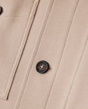 Close-up of a beige fabric from the Psycho Bunny MENS BEDFORD SHIRT JACKET - B6J290B200, showcasing a black button engraved with the logo and text, alongside parallel seams forming a rectangular pattern. The textured fabric suggests it's part of an item such as this shirt jacket.