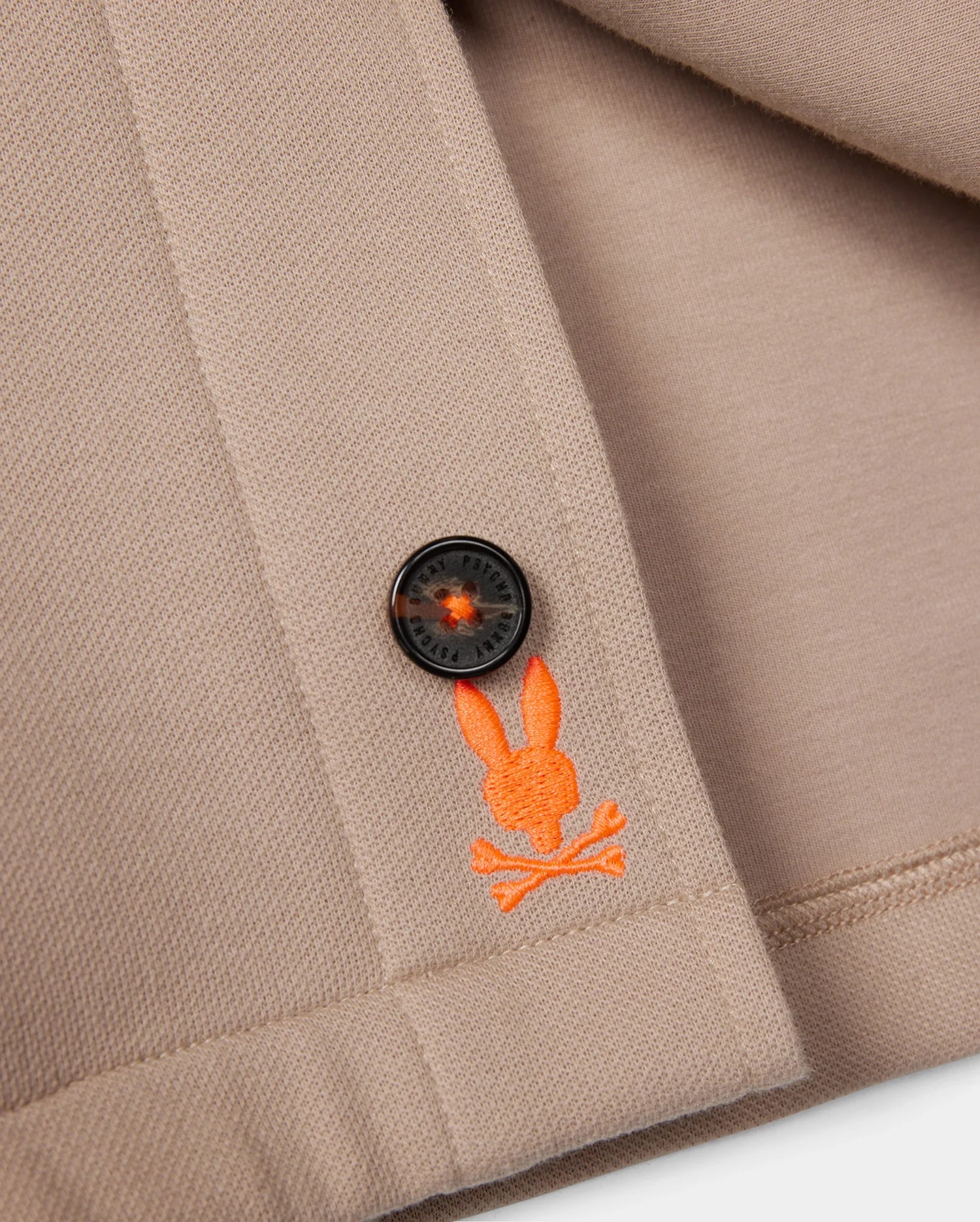 A close-up of the Psycho Bunny MENS BEDFORD SHIRT JACKET - B6J290B200 in beige showcases a minimalist design, featuring a logo-engraved black button and an orange embroidered emblem with a bunny head above crossed bones. The folded fabric highlights the detailed stitching, complemented by work-jacket style pockets.