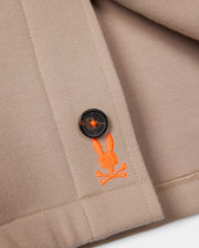 A close-up of the Psycho Bunny MENS BEDFORD SHIRT JACKET - B6J290B200 in beige showcases a minimalist design, featuring a logo-engraved black button and an orange embroidered emblem with a bunny head above crossed bones. The folded fabric highlights the detailed stitching, complemented by work-jacket style pockets.