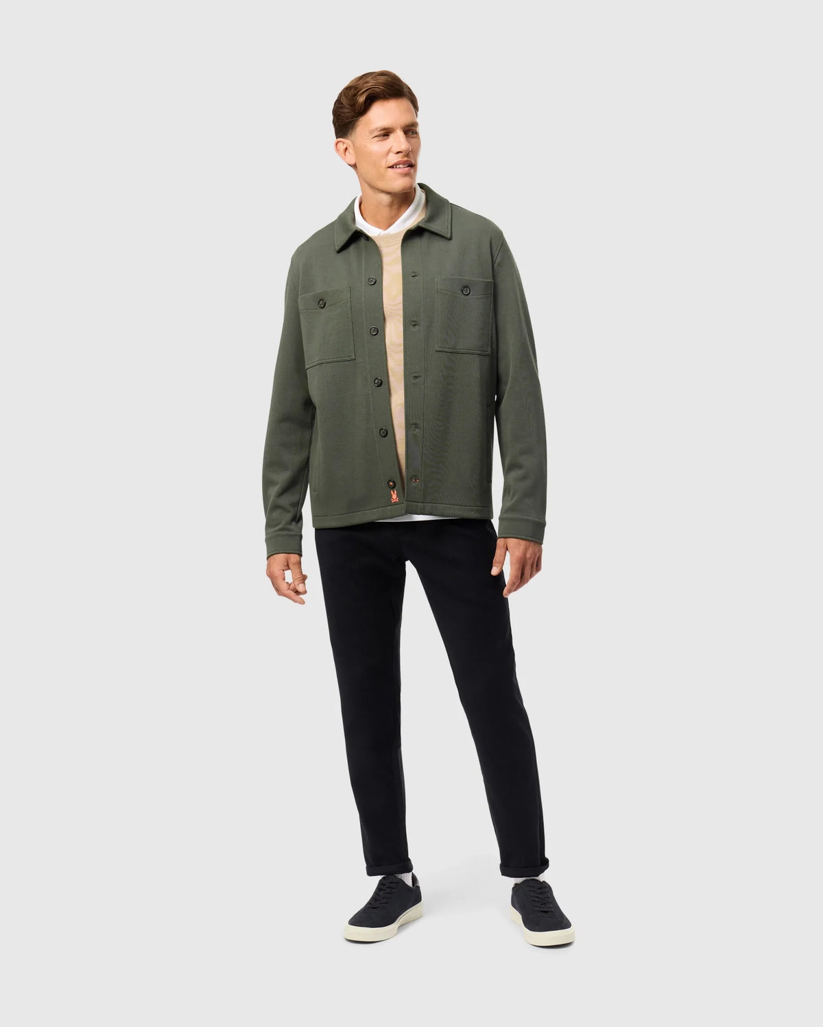 A man stands against a plain background, exuding an urban vibe in the Psycho Bunny MENS BEDFORD SHIRT JACKET - B6J290B200, styled over a beige cotton-blend shirt. He effortlessly pairs it with black pants and sneakers, while his short brown hair enhances his side glance and slight smile.