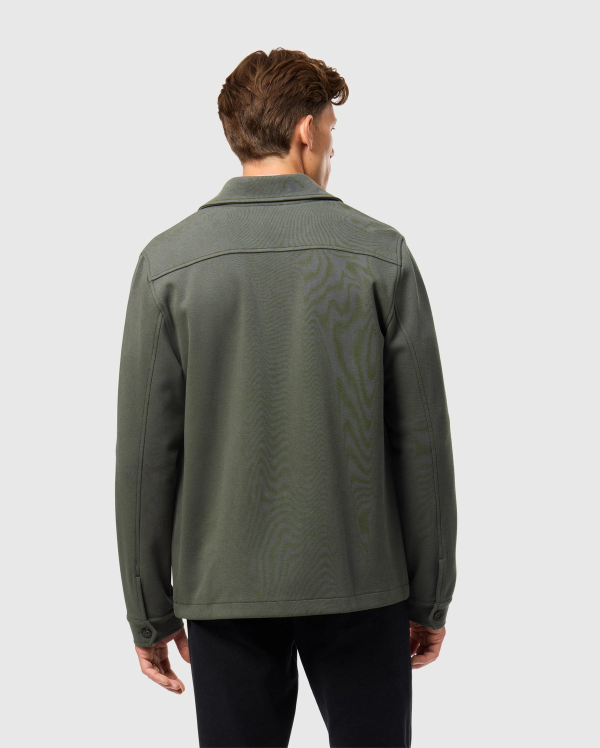 A person stands with their back to the camera, adorned in a Psycho Bunny MENS BEDFORD SHIRT JACKET - B6J290B200, paired with black pants. The outfit exudes an urban feel against a plain, light gray backdrop.
