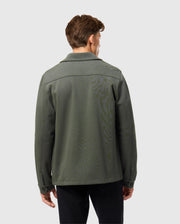 A person stands with their back to the camera, adorned in a Psycho Bunny MENS BEDFORD SHIRT JACKET - B6J290B200, paired with black pants. The outfit exudes an urban feel against a plain, light gray backdrop.