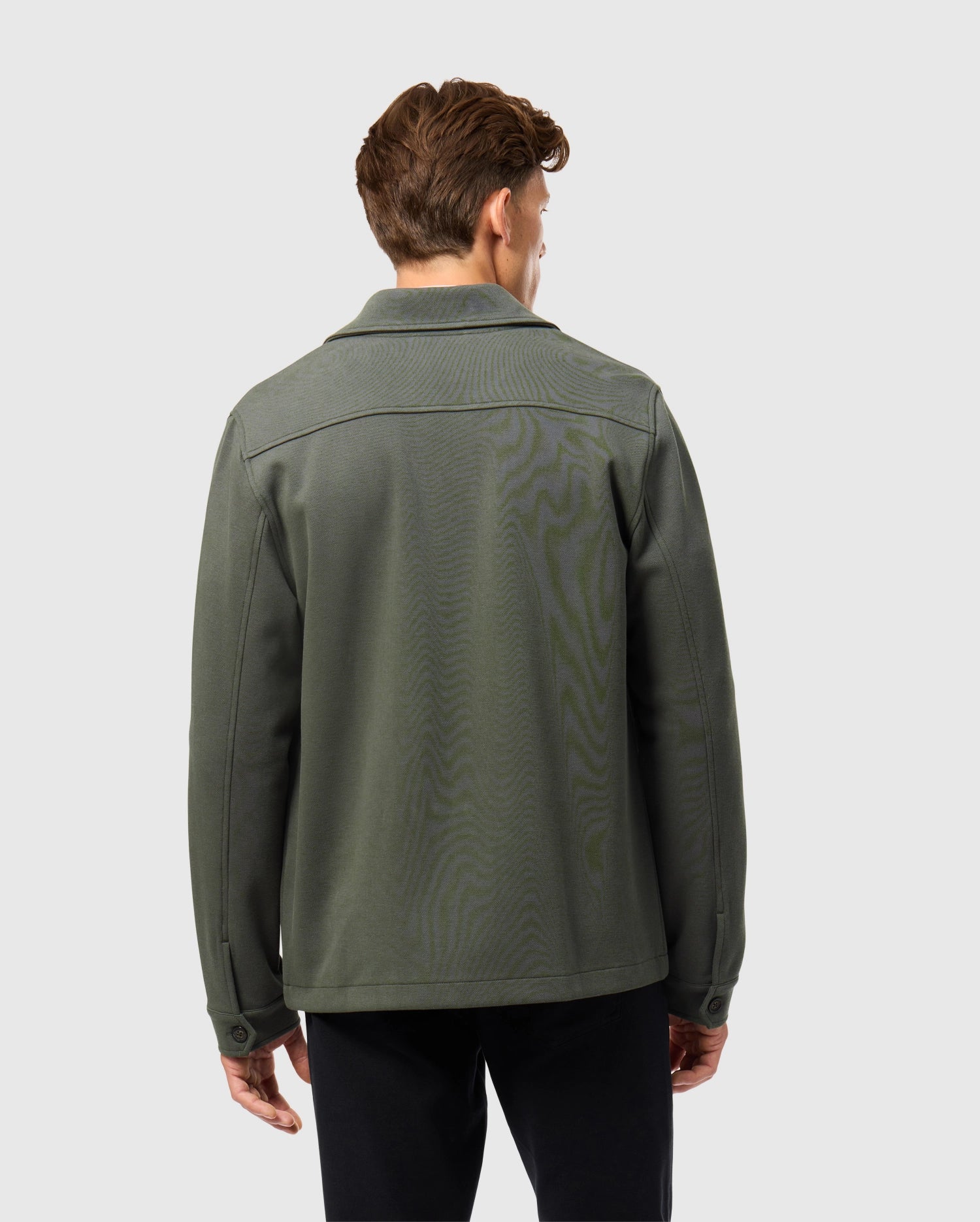 A person stands with their back to the camera, adorned in a Psycho Bunny MENS BEDFORD SHIRT JACKET - B6J290B200, paired with black pants. The outfit exudes an urban feel against a plain, light gray backdrop.