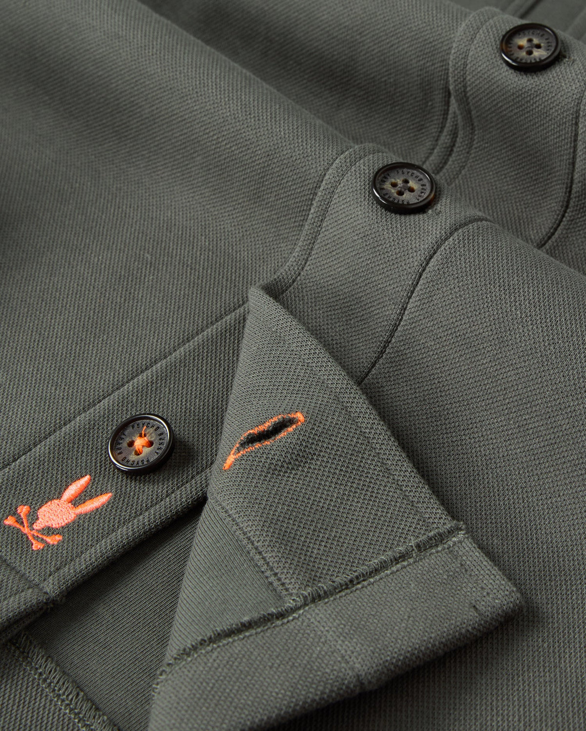 The "MENS BEDFORD SHIRT JACKET - B6J290B200" by Psycho Bunny features a close-up of green fabric adorned with black buttons and intricate orange embroidery depicting a rabbit alongside a small emblem. Its cotton-blend texture suggests an urban look, with the thickness and weaved appearance reminiscent of outerwear.
