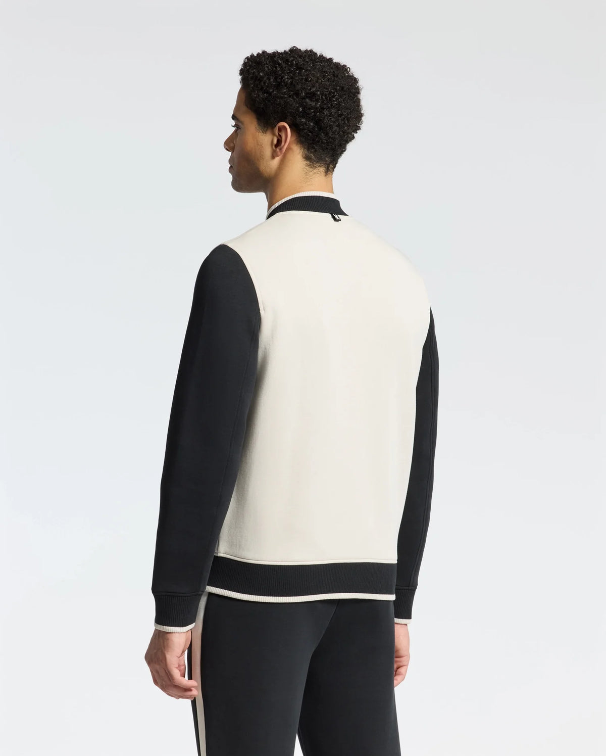 A person dons the Psycho Bunny MENS CRETE COLOR BLOCK BOMBER - B6J421F200 with black sleeves and a white back, paired with matching pants featuring a white stripe. The sleek silhouette stands against a plain background, facing away from the camera.