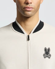 Wearing the Psycho Bunny MENS CRETE COLOR BLOCK BOMBER - B6J421F200, a person sports a classic light beige zip-up jacket with a black collar and stylized bunny emblem. The image captures them against a gray backdrop, face partially visible and cropped at the eyes.