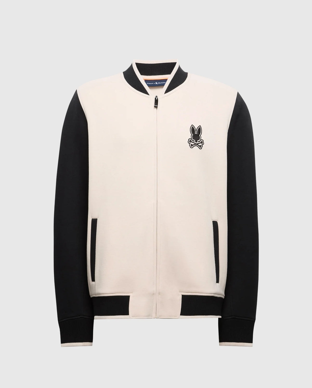 Introducing the Psycho Bunny MENS CRETE COLOR BLOCK BOMBER - B6J421F200: a varsity-style jacket with cream front, black sleeves, and trim. Featuring a rabbit crossbones logo on the chest, it's perfect for transitions with its zipper and side pockets.