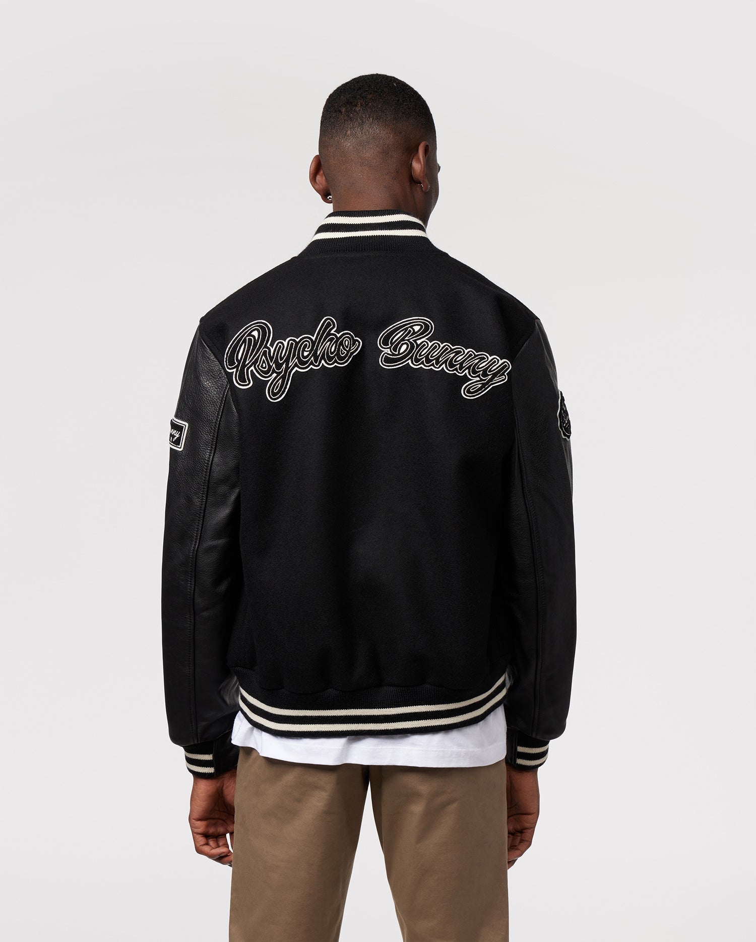 A person stands with their back to the camera, wearing the MENS ROBBINS VARSITY BOMBER JACKET - B6J524Z1OW by Psycho Bunny. The black jacket is adorned with chenille patches and features "Psycho Bunny" written across the back in white script. They also wear khaki pants and a white shirt beneath this stylish jacket.