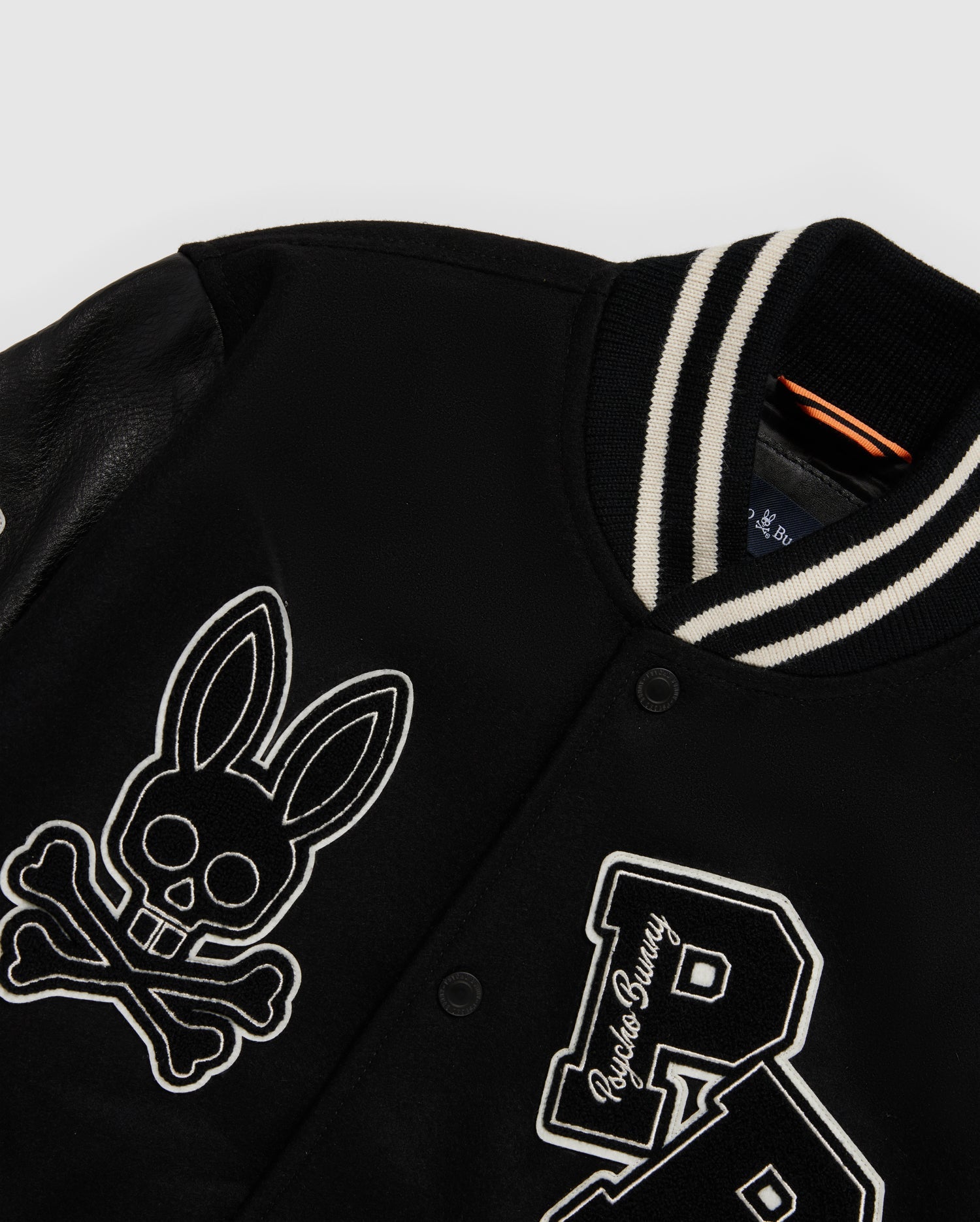 The MENS ROBBINS VARSITY BOMBER JACKET - B6J524Z1OW by Psycho Bunny is a black varsity jacket made from a cozy wool-blend, featuring leather sleeves and a striped collar. It is adorned with an eye-catching skull and crossbones symbol with bunny ears alongside a prominent "P," complete with standout chenille patches for added flair.