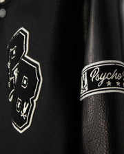 A detailed view of the MENS ROBBINS VARSITY BOMBER JACKET - B6J524Z1OW, crafted by Psycho Bunny. The black leather jacket is embellished with chenille patches, prominently displaying the number "23" with bunny designs. The sleeve features "Psycho" embroidered along with stars. The texture and elaborate stitching are remarkably evident.