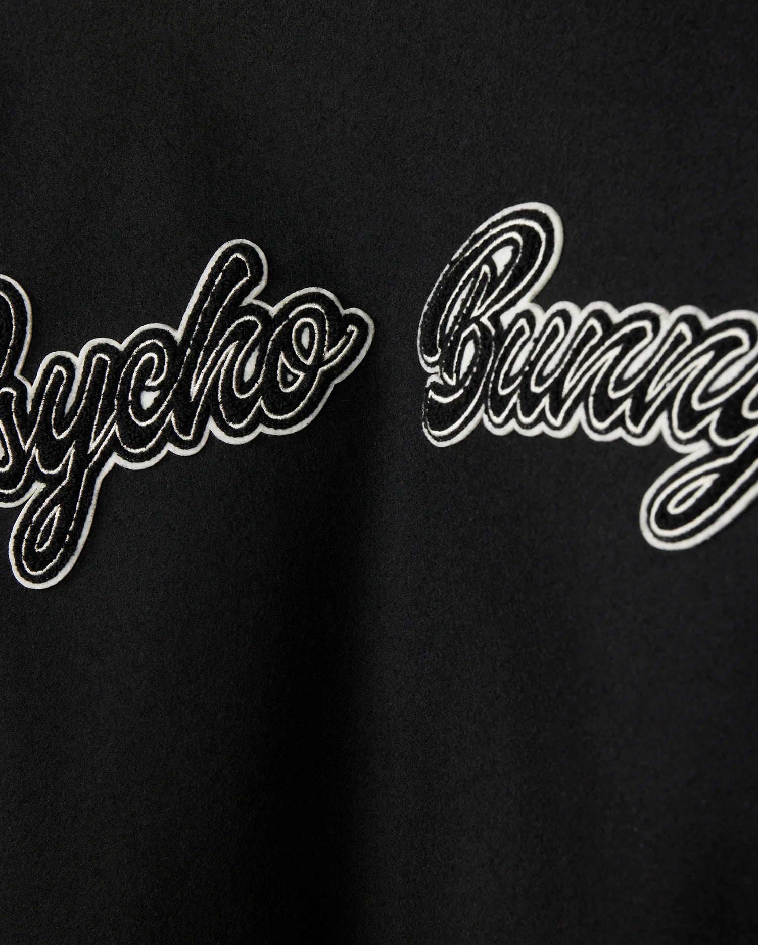 Close-up of a black fabric on the MENS ROBBINS VARSITY BOMBER JACKET (model B6J524Z1OW) by Psycho Bunny, showcasing the text "Psycho Bunny" in a cursive, embroidered style. The white text with a black outline contrasts against the wool-blend background, which is decorated with plush chenille patches.