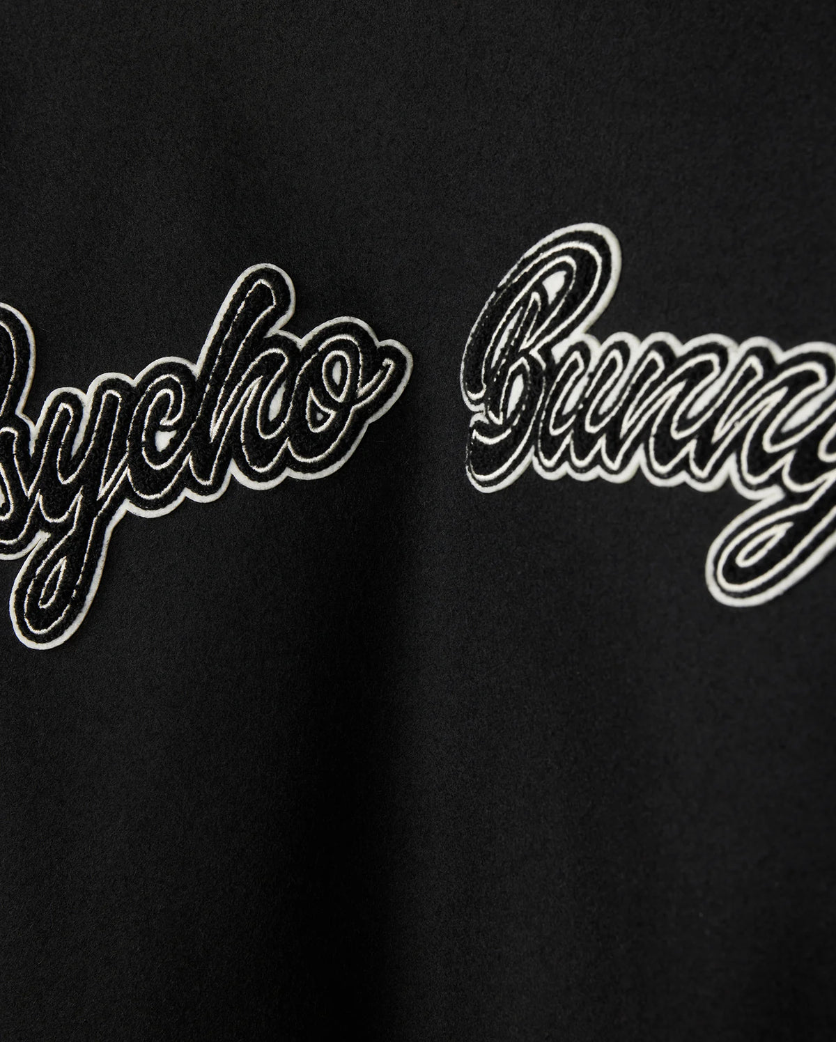 Close-up of a black fabric on the MENS ROBBINS VARSITY BOMBER JACKET (model B6J524Z1OW) by Psycho Bunny, showcasing the text "Psycho Bunny" in a cursive, embroidered style. The white text with a black outline contrasts against the wool-blend background, which is decorated with plush chenille patches.