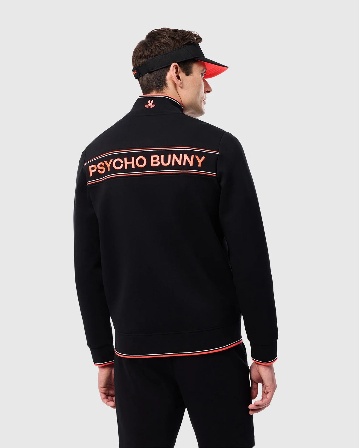 A man dressed in a black MENS COURTSIDE TRACK JACKET - B6J692C200 with orange trim and the words "Psycho Bunny" on the back. He is wearing a black visor with an orange brim. The background is plain and light gray.