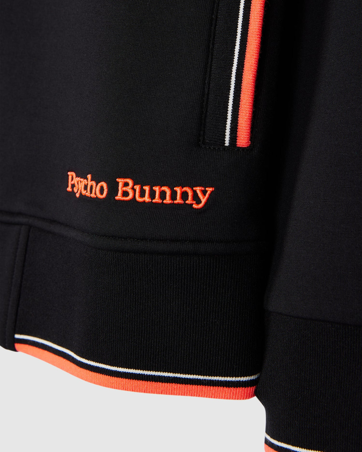 Close-up of a black Psycho Bunny MENS COURTSIDE TRACK JACKET - B6J692C200 with striped trim in orange, white, and black. The stretch cotton-blend jersey fabric features an embroidered "Psycho Bunny" logo in orange thread near the hem. The image highlights the texture, details of the stitching, and trim.