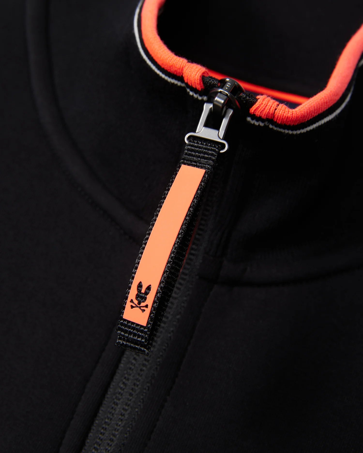 Close-up image of a black Psycho Bunny MENS COURTSIDE TRACK JACKET - B6J692C200 made from stretch cotton-blend jersey, featuring a bright orange zipper pull with a small skull and crossbones detail. The inner edge of the collar is also lined with bright orange, adding a pop of color to the garment.