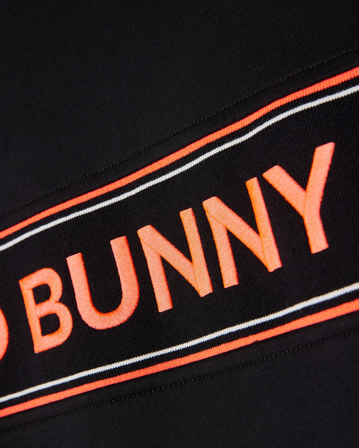 Close-up of a black **Psycho Bunny MENS COURTSIDE TRACK JACKET - B6J692C200** fabric with an orange and white stripe. The text "BUNNY" in bold orange letters is prominently displayed in the center of the stripe. The texture of the stretch cotton-blend jersey fabric is visible, giving it a detailed look.