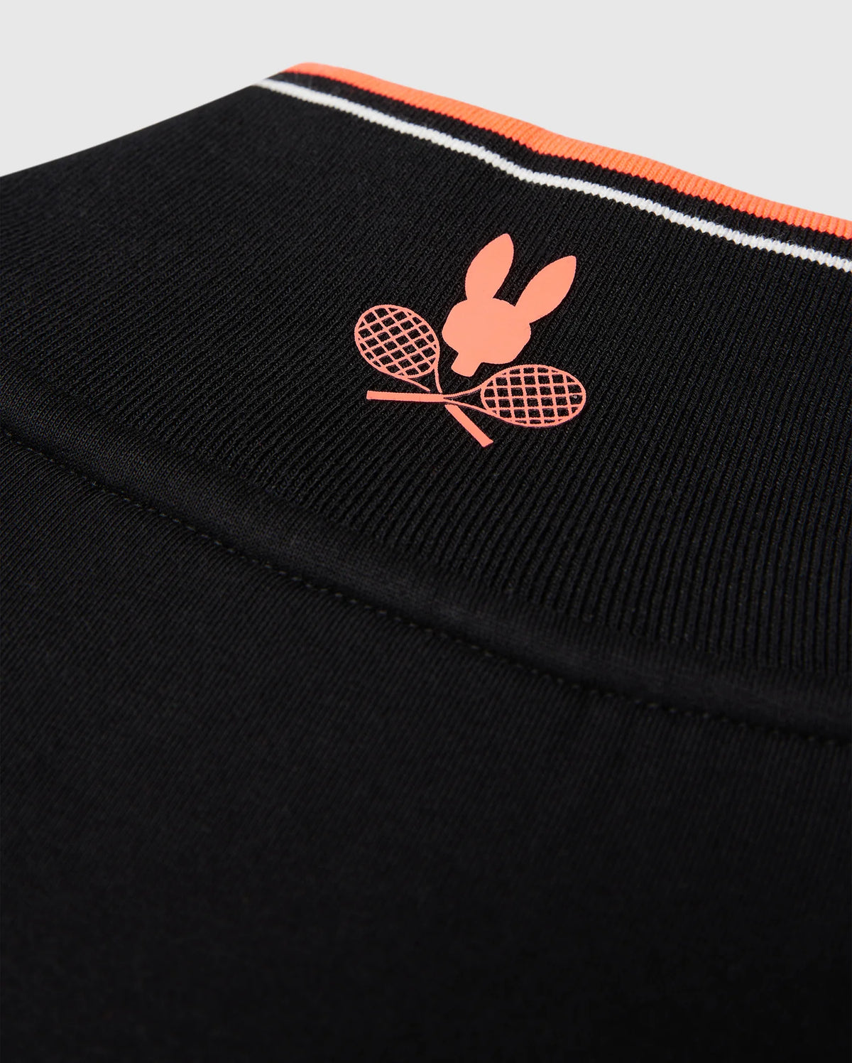 Close-up of a Psycho Bunny MENS COURTSIDE TRACK JACKET - B6J692C200 made from stretch cotton-blend jersey, featuring a logo with a red silhouette of a rabbit's head above two crossed red tennis rackets. The garment has a thin red, white, and black striped band near the top edge and logo-engraved hardware.