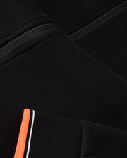 Close-up of a black fabric garment featuring a seam detail and an orange stripe with white and black accents. The image highlights the texture, design elements of the textile, and logo-engraved hardware typical of a Psycho Bunny MENS COURTSIDE TRACK JACKET - B6J692C200.