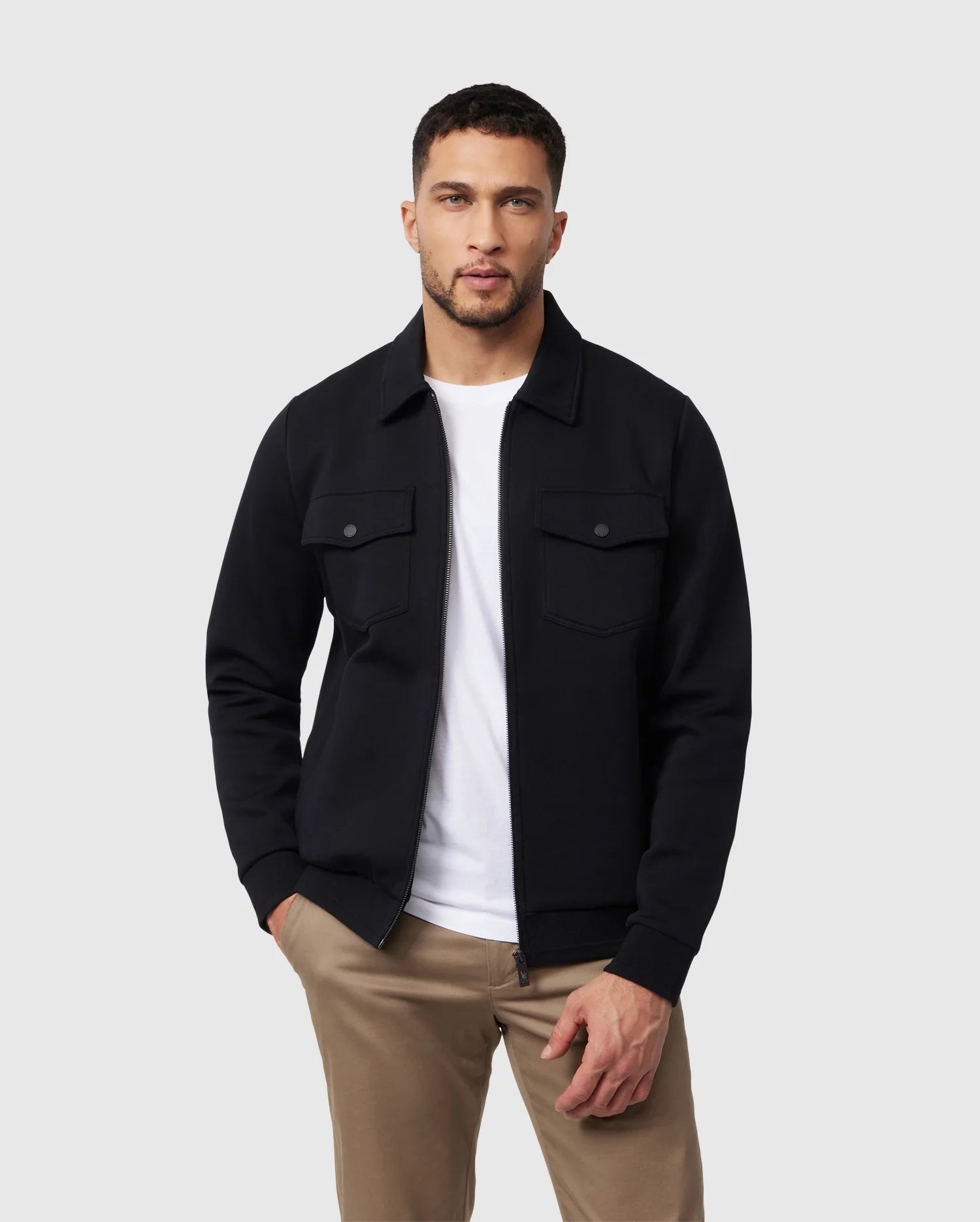 A man stands against a light grey background wearing the Psycho Bunny Men's Oroville Bomber Jacket (B6J712A200) in black over a white T-shirt and beige pants. With his hands in his pockets, short hair, and a beard, he effortlessly showcases how this outerwear seamlessly blends style with comfort.