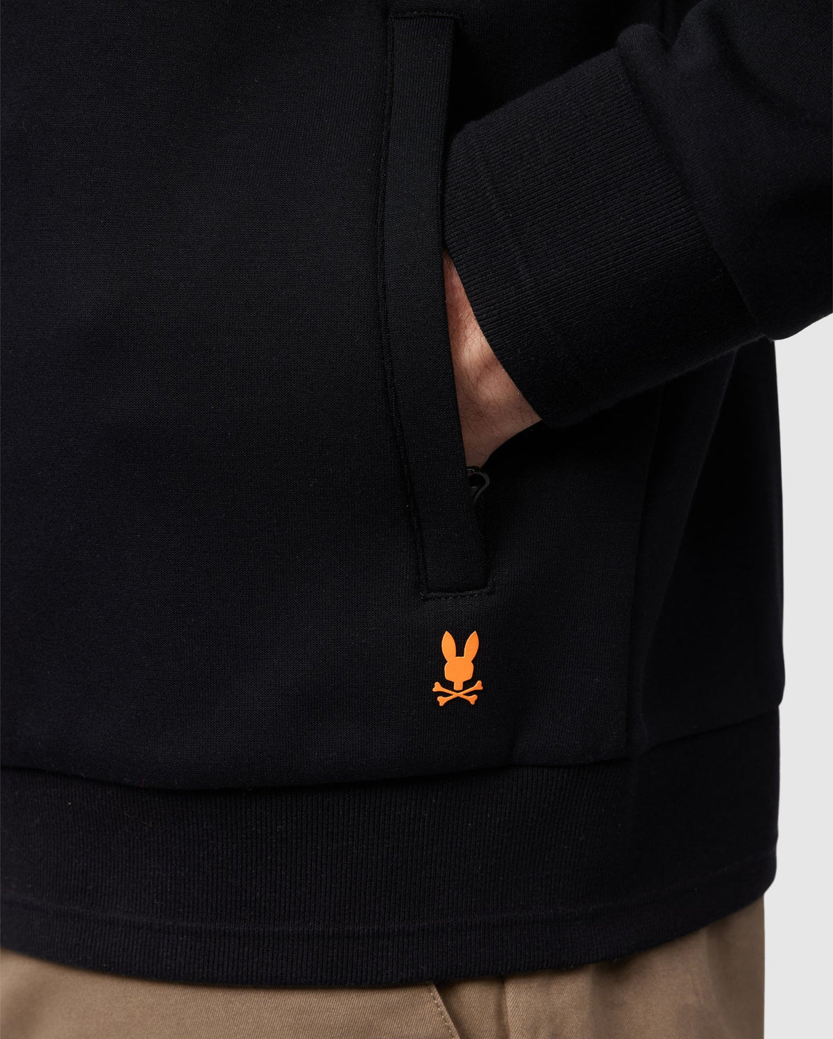 A person is wearing the MENS OROVILLE BOMBER JACKET - B6J712A200 by Psycho Bunny in black, casually tucking one hand into their pocket. This cotton-poly blend jacket stands out with its striking orange bunny with crossbones logo near the hem. They are also styled with beige pants to complete the look.