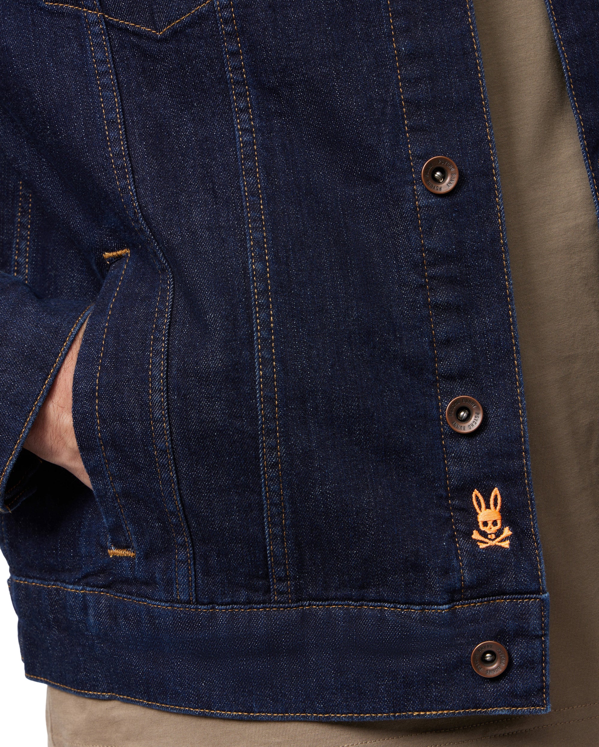 A close-up of someone wearing the MENS PATHE DENIM JACKET - B6J741X1CO by Psycho Bunny, characterized by its raw denim fabric and brown buttons that define men's fashion. The jacket is adorned with an orange embroidered rabbit logo above crossed bones on the lower right side. One hand is casually resting in the pocket, enhancing the ensemble with a brown shirt.