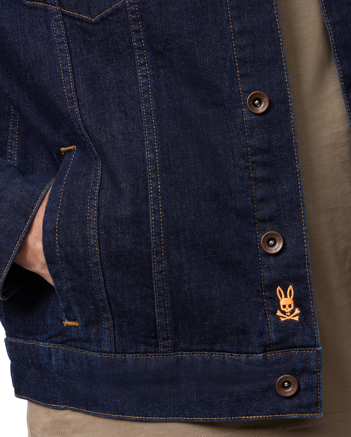 A close-up of someone wearing the MENS PATHE DENIM JACKET - B6J741X1CO by Psycho Bunny, characterized by its raw denim fabric and brown buttons that define men's fashion. The jacket is adorned with an orange embroidered rabbit logo above crossed bones on the lower right side. One hand is casually resting in the pocket, enhancing the ensemble with a brown shirt.