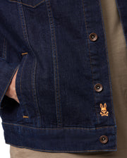 A close-up of someone wearing the MENS PATHE DENIM JACKET - B6J741X1CO by Psycho Bunny, characterized by its raw denim fabric and brown buttons that define men's fashion. The jacket is adorned with an orange embroidered rabbit logo above crossed bones on the lower right side. One hand is casually resting in the pocket, enhancing the ensemble with a brown shirt.