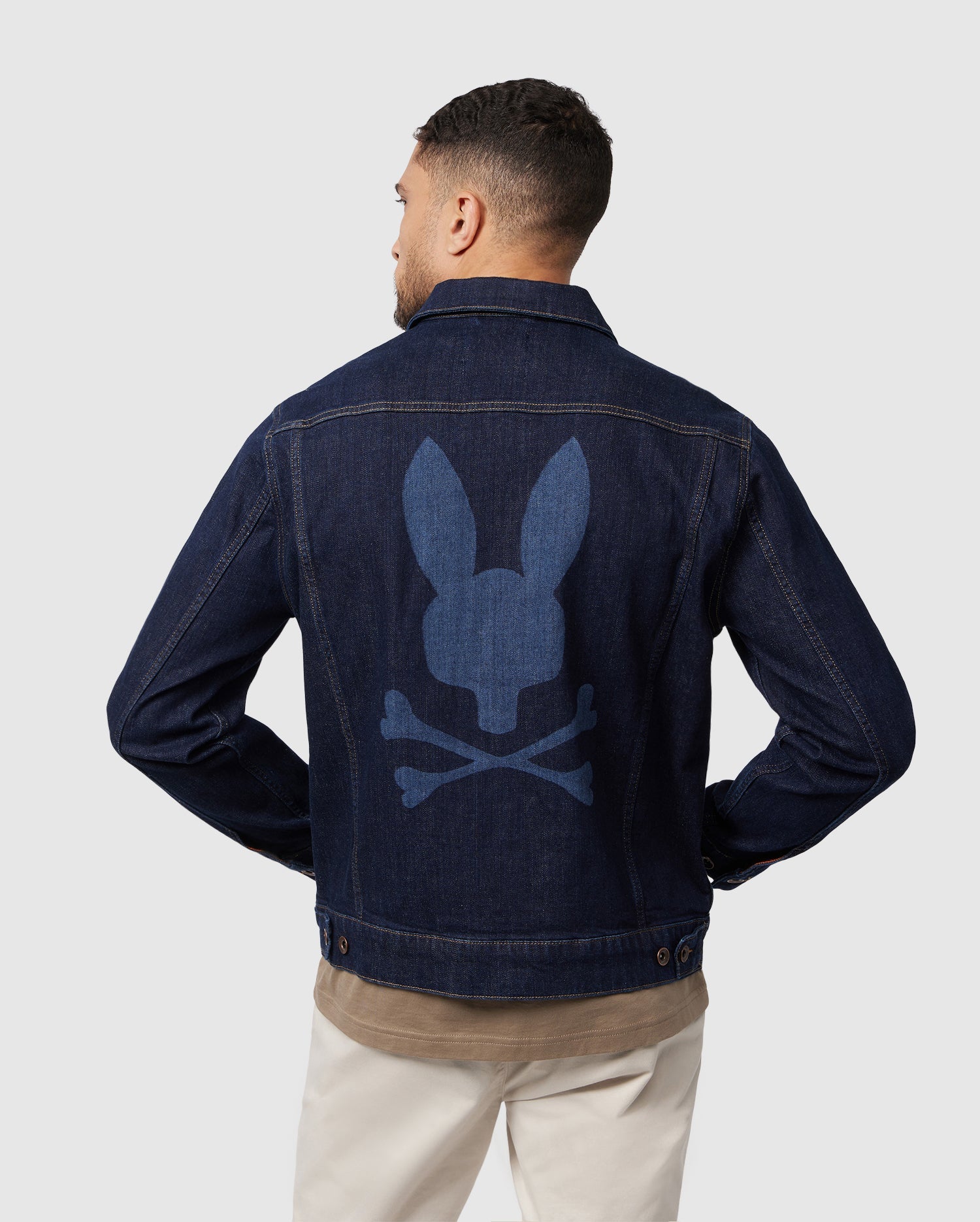 A man sports the Psycho Bunny MENS PATHE DENIM JACKET - B6J741X1CO, featuring an embroidered bunny skull and crossbones design on the back. With his back to the camera, he effortlessly pairs it with a brown shirt and beige pants against a plain background, exuding coolness.