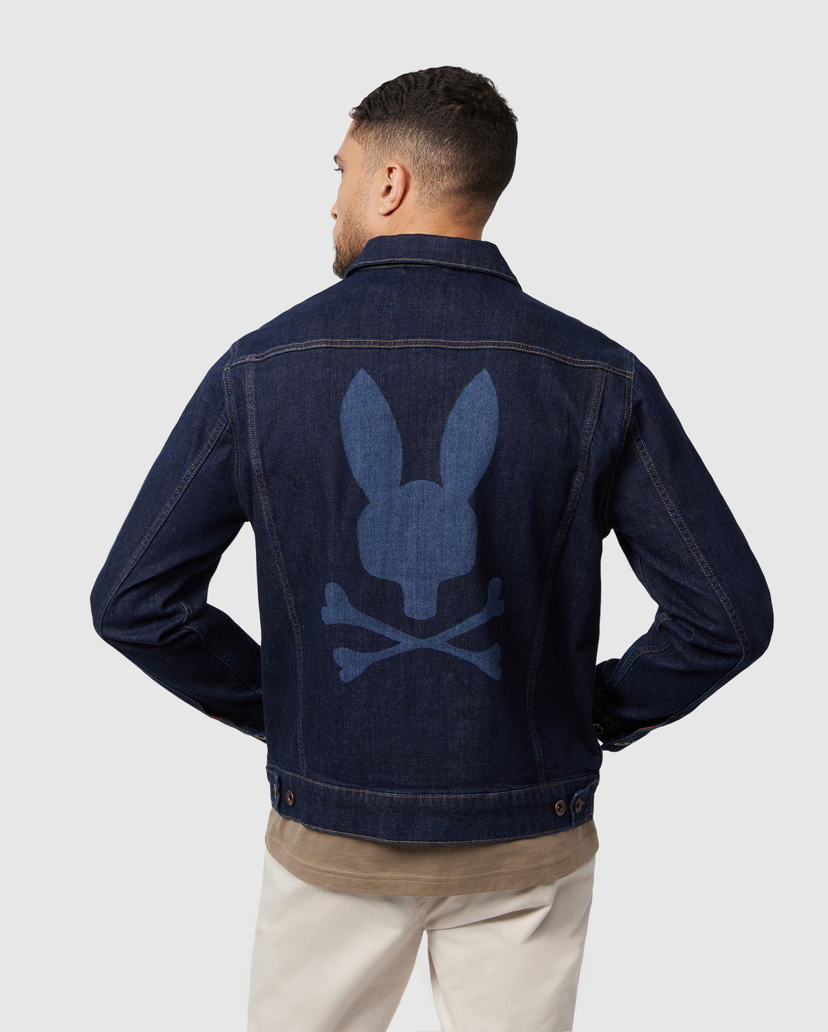 A man sports the Psycho Bunny MENS PATHE DENIM JACKET - B6J741X1CO, featuring an embroidered bunny skull and crossbones design on the back. With his back to the camera, he effortlessly pairs it with a brown shirt and beige pants against a plain background, exuding coolness.