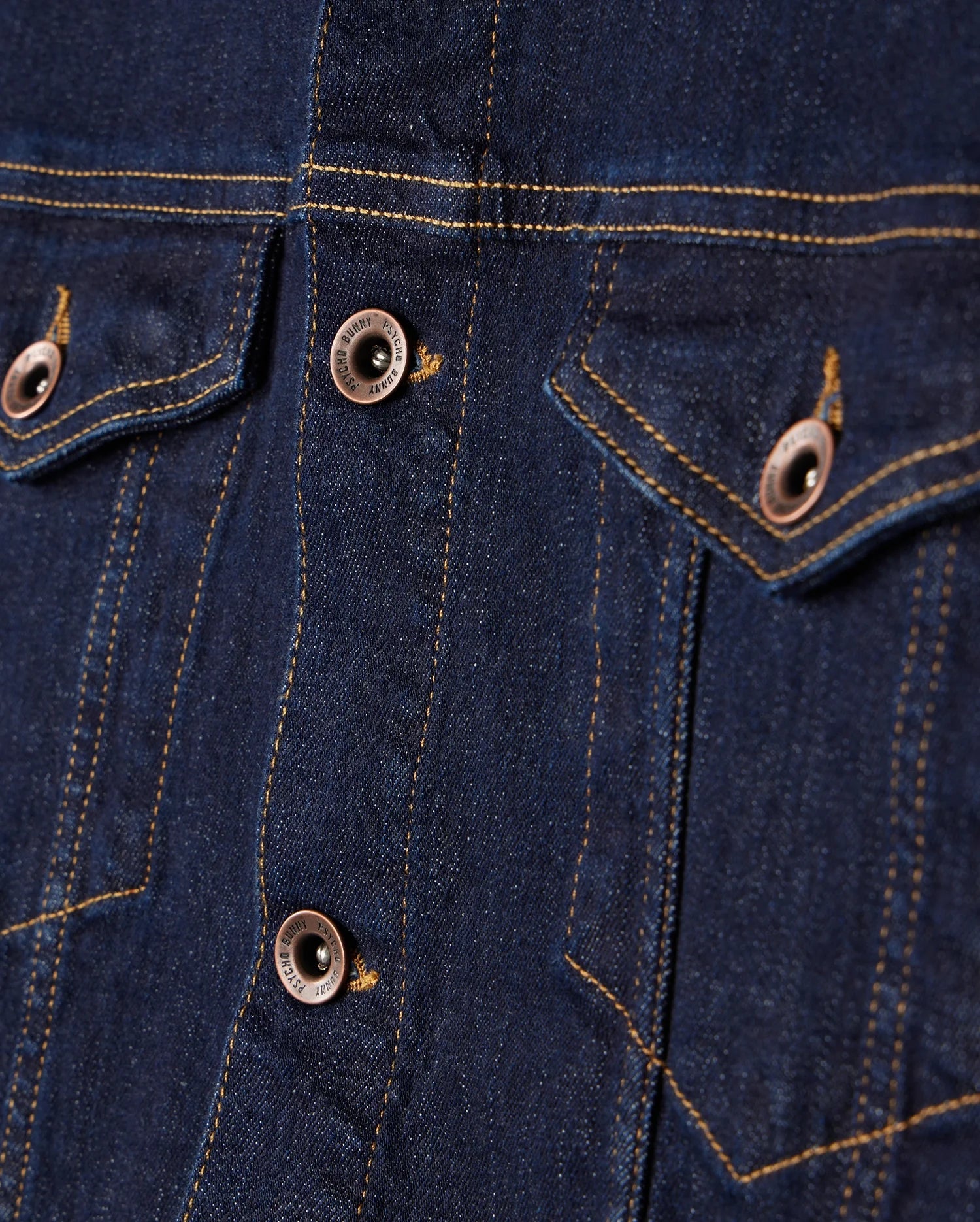 A close-up of the MENS PATHE DENIM JACKET - B6J741X1CO by Psycho Bunny highlights its raw denim finish with two buttoned chest pockets featuring contrasting stitching. The blue fabric and metal buttons enhance this classic design in men's fashion.