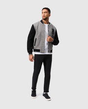 A man stands wearing the Psycho Bunny MENS TOM WOOL BOMBER JACKET - B6J744D200, which features black sleeves and a sophisticated design. He pairs it with a white T-shirt, black pants, and black sneakers. The jacket is adorned with a distinctive black emblem on the left side. He gazes to his left against a simple light gray background.