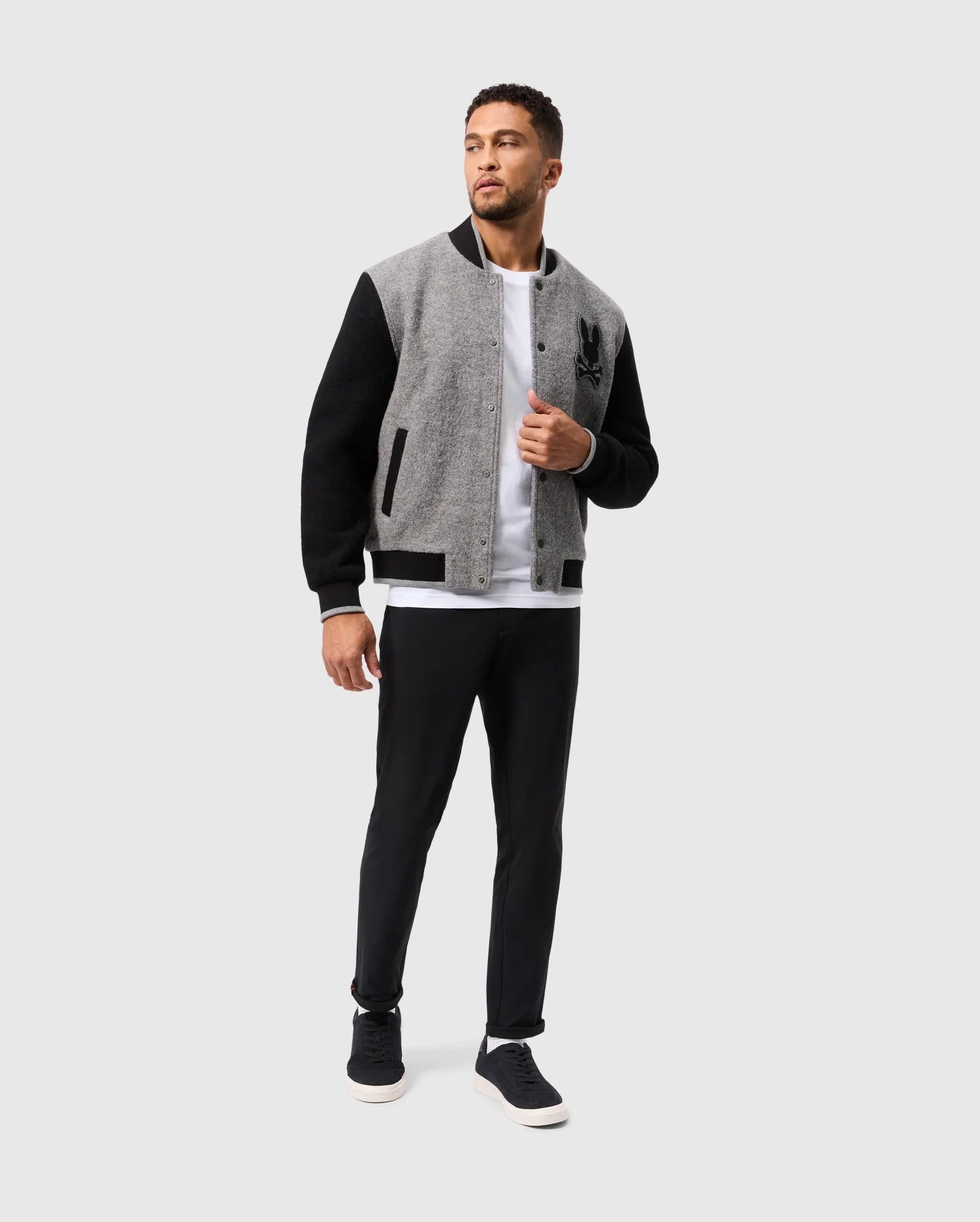 A man stands wearing the Psycho Bunny MENS TOM WOOL BOMBER JACKET - B6J744D200, which features black sleeves and a sophisticated design. He pairs it with a white T-shirt, black pants, and black sneakers. The jacket is adorned with a distinctive black emblem on the left side. He gazes to his left against a simple light gray background.