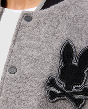 Close-up of the stylish Men's Tom Wool Bomber Jacket by Psycho Bunny, featuring a gray design adorned with a black rabbit and crossbones patch on the side. The jacket, identified as product B6J744D200, showcases a round black button with part of a white shirt peeking out from beneath.