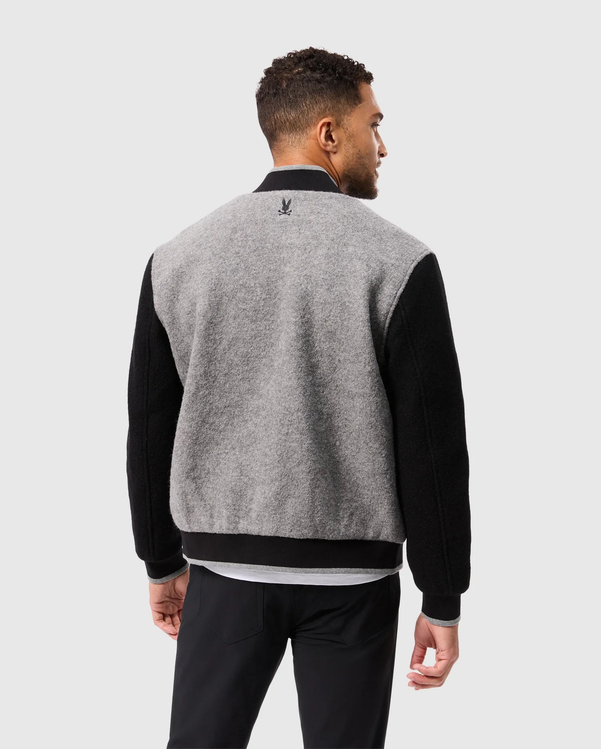 A man wearing the MENS TOM WOOL BOMBER JACKET - B6J744D200 by Psycho Bunny, featuring an elegant gray and black design with a subtle emblem on the back, stands facing away. He has short curly hair styled in a casual manner and pairs it with black pants. The backdrop is a plain light gray.