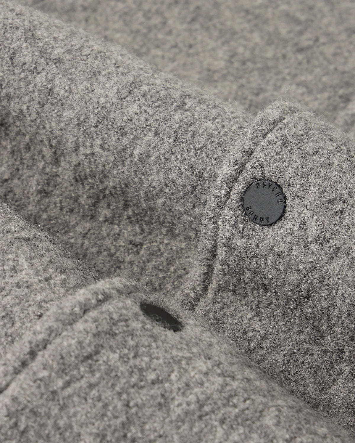 Close-up of a grey woolen fabric with a textured surface reminiscent of the MENS TOM WOOL BOMBER JACKET - B6J744D200 by Psycho Bunny. The fabric is adorned with a black button embossed with text, which provides a subtle contrast against the soft material.