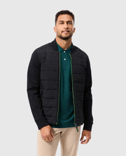 A man with short curly hair is wearing the Psycho Bunny Men's Mykel Mixed Media Jacket in black, layered over a green polo shirt and beige pants. He stands against a plain light gray background, gazing at the camera with a slight smile.