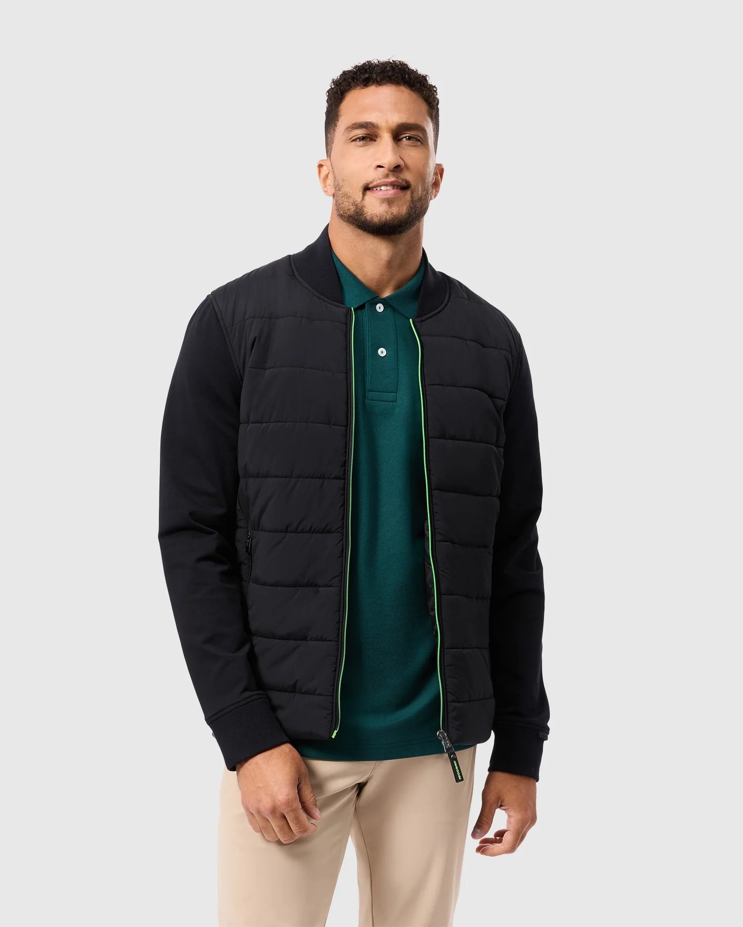 A man with short curly hair is wearing the Psycho Bunny Men's Mykel Mixed Media Jacket in black, layered over a green polo shirt and beige pants. He stands against a plain light gray background, gazing at the camera with a slight smile.