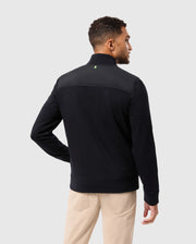 A person with short hair is wearing a versatile Psycho Bunny MENS MYKEL MIXED MEDIA JACKET in black and beige pants, standing with their back to the camera against a light gray background.