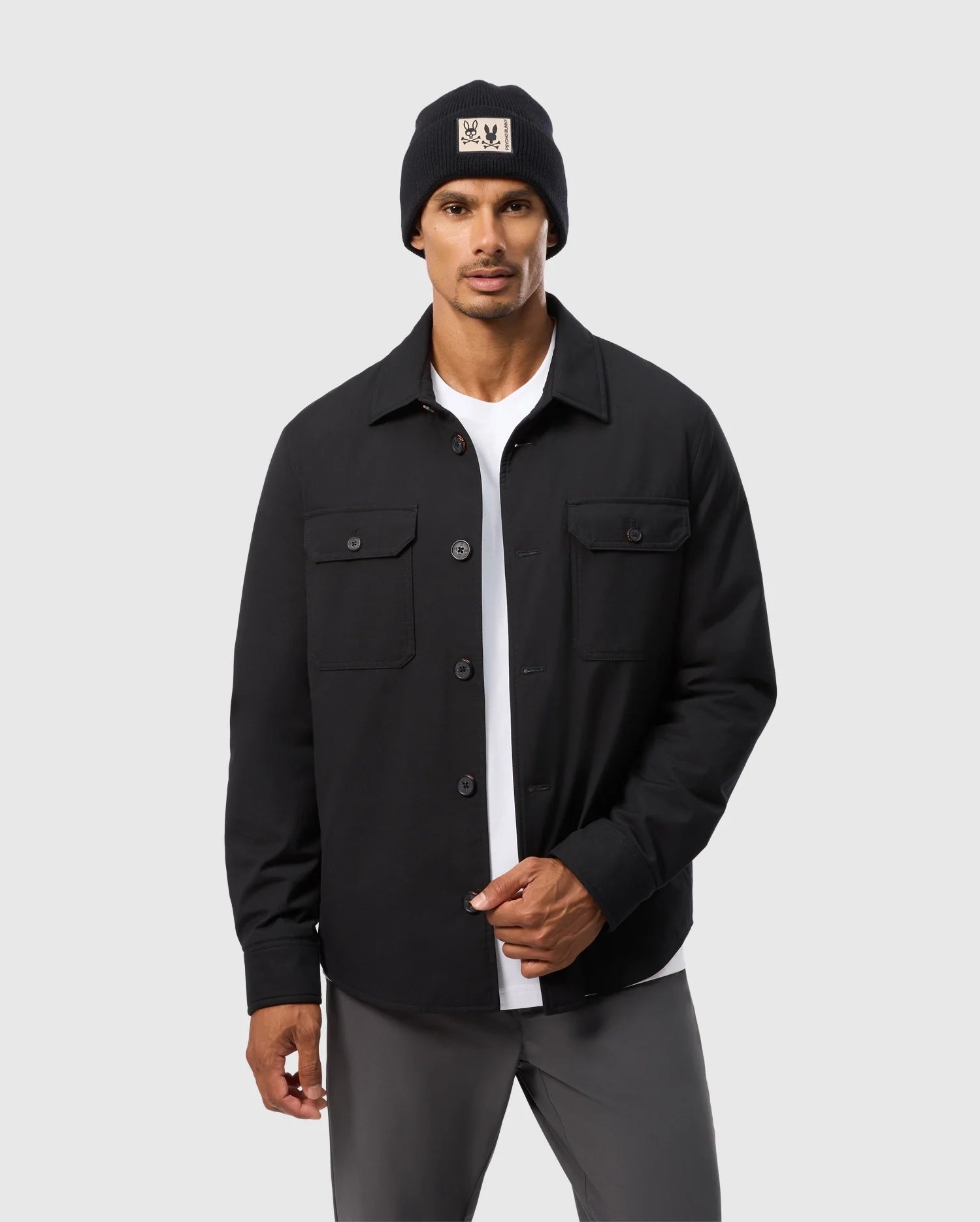 A man sports a Psycho Bunny MENS HILLCREST PADDED SHACKET (B6J936D200) with subtle neon lining over a white shirt and gray pants, topped off with a black beanie, against a plain gray background.