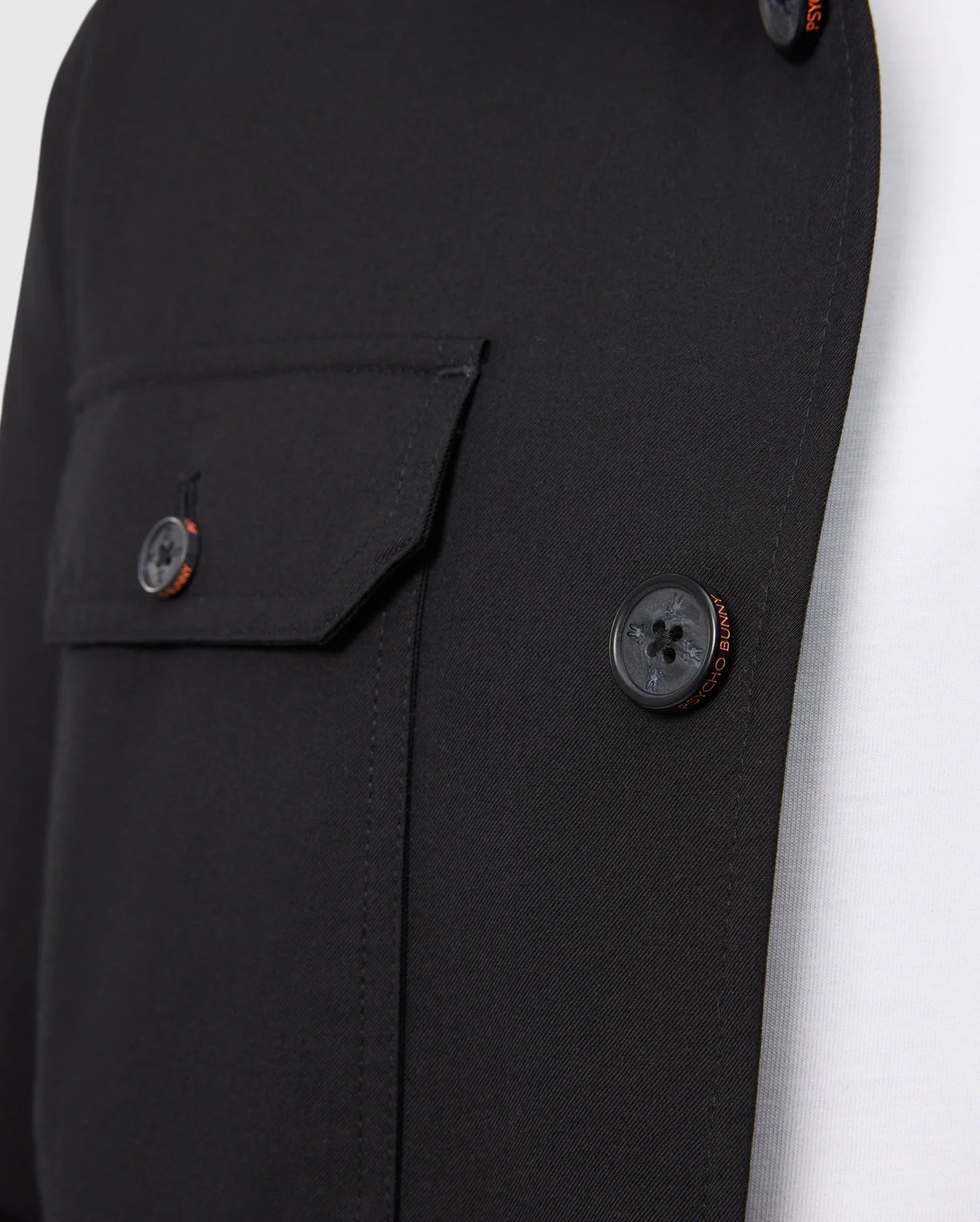A close-up of the Psycho Bunny MENS HILLCREST PADDED SHACKET - B6J936D200, in black, showing visible buttons and a left flap pocket, layered over a sleek white shirt.