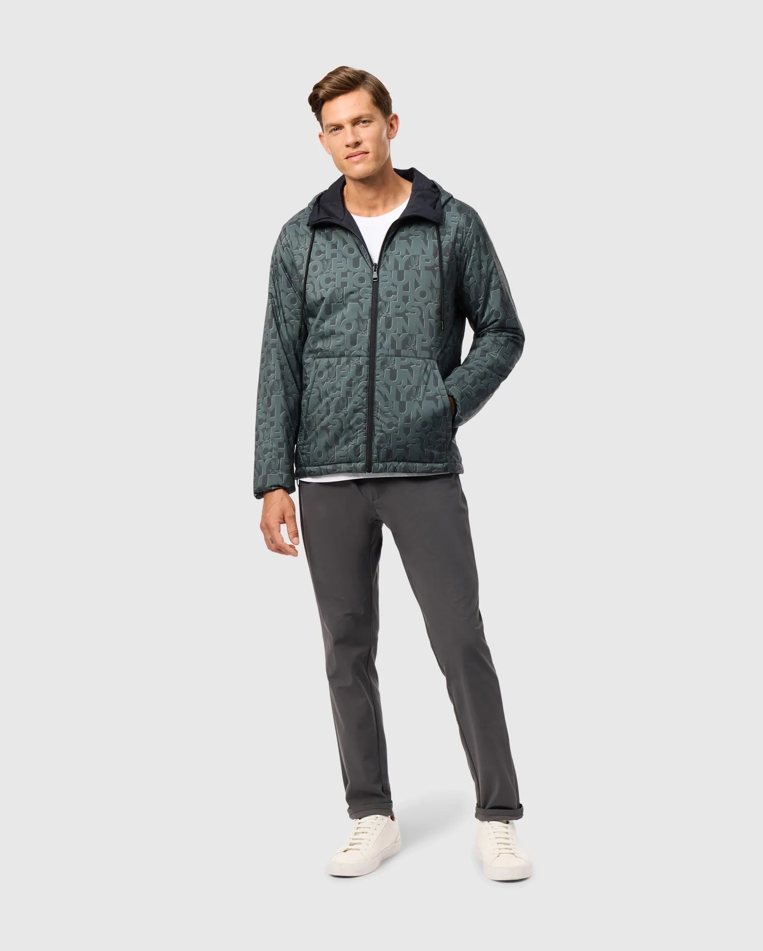A man in a Psycho Bunny SOUTHWORTH REVERSIBLE PADDED HOODED JACKET (B6J946D200), wearing a white t-shirt, dark gray pants, and white sneakers, stands against a light gray background. One hand is in his jacket pocket as he maintains a neutral expression.